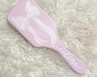 May include: A pink hairbrush with a white bow painted on the back and the name "celine" written on the handle.