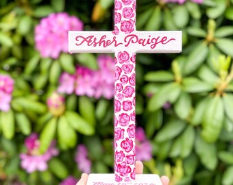 May include: White wooden cross with a pink floral design. The cross has the names "Asher Paige" and the date "May 18th 2024" written on it.