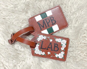 May include: Two brown leather luggage tags. One tag features the initials "MPB" in black with white and green accents. The other tag has the initials "LAB" in black, surrounded by a white floral design. Both tags have brown leather straps.