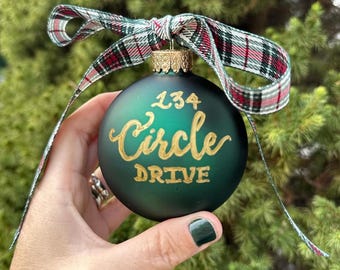 May include: A green glass Christmas bauble with gold lettering that reads "134 Circle Drive". The bauble is tied with a red and white tartan ribbon.