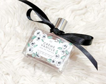 May include: A clear perfume bottle with a silver cap and a black ribbon. The label reads "Crème Vanille Eau de Parfum" with floral accents. The bottle contains 30 ml of perfume.