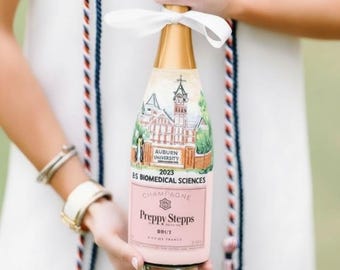 May include: A champagne bottle with a gold top and a pink label. The label reads "Preppy Stepps Brut" and "Auburn University 2023 BS Biomedical Sciences". The bottle is tied with a white ribbon.