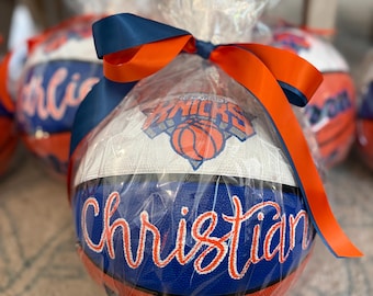 May include: A basketball featuring the New York Knicks logo and the name Christian written in orange script, wrapped in clear plastic with a blue and orange satin ribbon tied at the top. The ball has blue and orange panels with white accents.