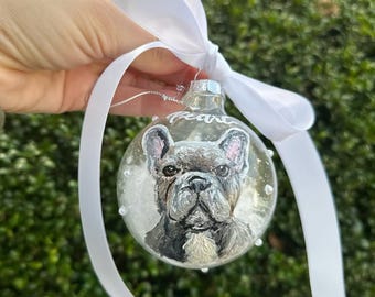 May include: A clear glass Christmas bauble with a hand-painted portrait of a French Bulldog. The bauble is decorated with white glitter and the name "Pearce" is written on the front.