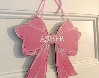 May include: A pink wooden bow-shaped sign with the name "ASHER" in white lettering. The sign is hanging on a white door with a pink ribbon. The bow has a black outline.