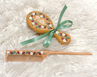 May include: A wooden hairbrush and comb set featuring a strawberry design. The oval brush is inscribed with "Pieta". Both items are decorated with red strawberries, white flowers, and green leaves, and are tied with a green ribbon.