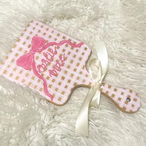 May include: A pink and gold checkered wooden cutting board with a pink bow and the name "Carlie Mae" written in cursive. A cream-colored ribbon is tied around the handle.