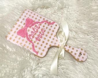 May include: A pink and gold checkered wooden cutting board with a pink bow and the name "Carlie Mae" written in cursive. A cream-coloured ribbon is tied around the handle.