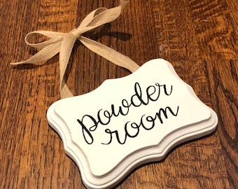 May include: White wooden sign with a brown ribbon attached. The sign has a scalloped edge and says "Powder Room" in black cursive lettering.