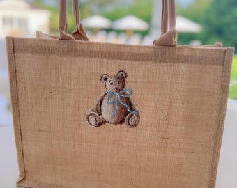 May include: A brown burlap tote bag with a brown teddy bear with a blue bow embroidered on the front.