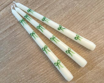 May include: Three white taper candles with green bows printed on them. The candles are arranged in a row on a wooden surface.