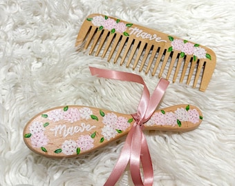 May include: A wooden comb and brush set, decorated with pink and white floral patterns and the name "Maeve". A pink satin ribbon ties the brush. The comb has a handle and the brush is oval-shaped.