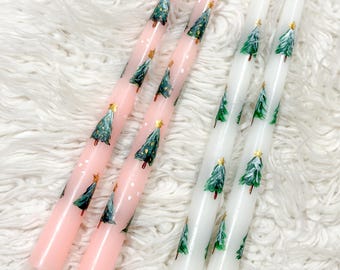 May include: Pink and white taper candles decorated with green Christmas trees and gold star accents. The candles are arranged on a white, fluffy surface, creating a festive holiday theme.