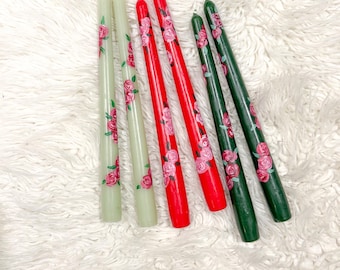 May include: Six taper candles in shades of green, red, and dark green, decorated with pink rose floral patterns. The candles are arranged on a white, fluffy surface.