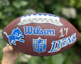 May include: A brown leather American football featuring white laces, a blue Detroit Lions logo, the text Wilson, the number 17, the word LIONS in blue and white, and a central NFL shield emblem with blue and white detailing on the textured surface.