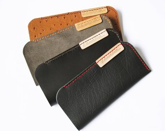 May include: Three leather phone cases in different colors and textures. The top case is brown with a textured pattern, the middle case is gray, and the bottom case is black. Each case has a small, rectangular label with the text "Leather Goods" printed on it.