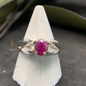 May include: A silver ring featuring a pink oval gemstone flanked by two smaller clear gemstones.