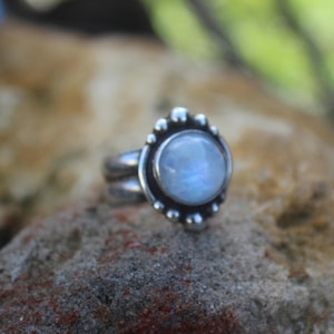 May include: A silver ring featuring a round, translucent, iridescent moonstone cabochon set in a bezel. The stone is surrounded by a decorative border of small, polished silver spheres, and the band is constructed from two parallel silver wires.