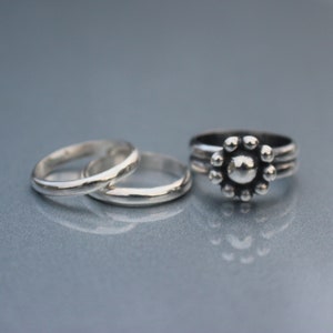 May include: Three silver rings are displayed on a reflective surface. Two are simple bands, while the third features a floral design with a central sphere surrounded by smaller spheres. The rings are likely made of sterling silver.