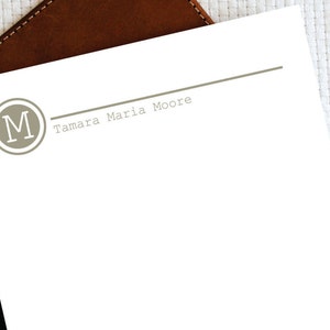 Può includere: Un foglio di carta bianco con una linea grigia e il testo "Tamara Maria Moore" stampato su di esso. Un cerchio grigio con la lettera "M" in bianco si trova nell'angolo in alto a sinistra.