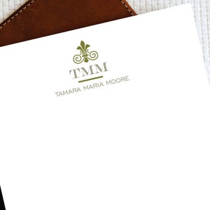Könnte beinhalten: Eine weiße Briefpapierkarte mit einem grauen und grünen Logo. Das Logo zeigt eine stilisierte Fleur-de-lis und den Text "TMM" über dem Namen "TAMARA MARIA MOORE".
