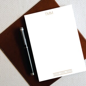 Puede incluir: Un bloc de notas blanco con las iniciales "TMM" impresas en dorado en la parte superior y el nombre "TAMARA MARIA MOORE" impreso en dorado en la parte inferior. Un bol&iacute;grafo negro est&aacute; sobre el bloc de notas.