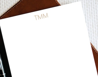 Op de afbeelding: Een wit notitieboekje met de letters "TMM" in bruin gedrukt bovenaan de pagina. Het notitieboekje ligt op een bruin lederen oppervlak.