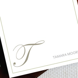 Może przedstawiać: Biały notes ze zieloną ramką i szarym, ozdobnym literą "T" w środku. Na dole notesu znajduje się napis "TAMARA MOORE" w kolorze szarym.