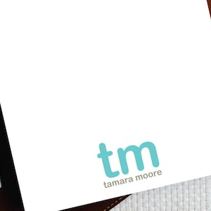 May include: A white notepad with a brown leather cover and a blue and white logo that says "tm tamara moore".