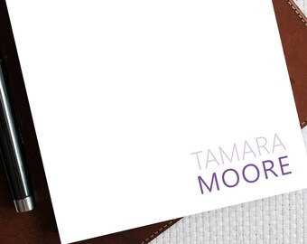 May include: A white notepad with the name "TAMARA MOORE" printed in purple and light purple on the bottom right corner. A black pen is lying on a brown leather surface next to the notepad.