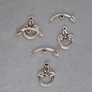 May include: Four silver-toned metal clasps with a floral design. The clasps are in the shape of a circle with a leaf design on each side. Two of the clasps have a curved bar attached to the top.