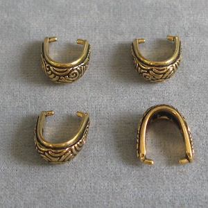 May include: Four gold-colored jewelry findings, each with a U-shaped design. The findings have an ornate, engraved pattern on the front and sides. The background is a light gray fabric.