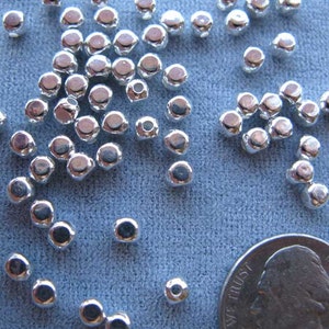 May include: A close-up of a pile of silver-colored metal beads, with a US quarter for scale. The beads are round and have a slightly domed shape.