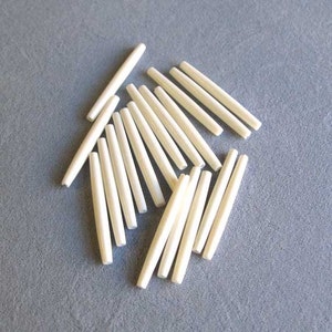 May include: A group of 16 white glass beads, each shaped like a long, thin cylinder. The beads are scattered on a gray surface.
