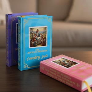 May include: Three hardback books are displayed on a dark wooden surface. The books are in shades of blue, purple, and pink. The blue book has the text "The Book of Mormon" and "Ceardry Smith". The pink book has the text "Book of Mormon, Doctrine and Covenants, Pearl of Great Price".