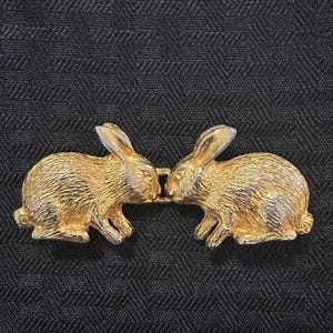 May include: A gold-toned metal belt buckle featuring two rabbits facing each other. The rabbits have detailed, textured fur patterns and are connected in the center by a small rectangular link. The design captures the rabbits in a seated position.