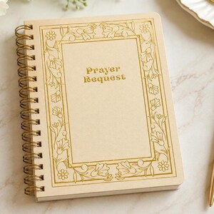 May include: A spiral-bound notebook with a light beige cover featuring a gold foil floral border and the text Prayer Request printed in the center. The notebook has a gold metal wire coil binding along the left side and a rectangular shape.