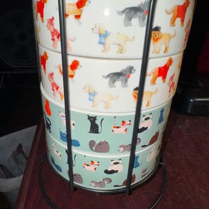 May include: A set of four stacked ceramic bowls held in a black metal wire stand. The top two bowls are white with a pattern of dogs wearing scarves and sweaters. The bottom two bowls are light green with a pattern of various cats in different poses.