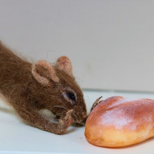 May include: A brown and white felted mouse figurine looking at a heart-shaped bread roll.