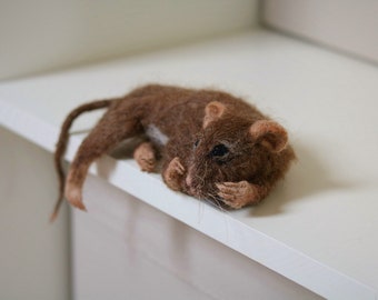 May include: A brown and white needle felted mouse with a long tail, lying on a white shelf.