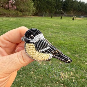 Peut inclure: Une broche en forme d'oiseau réalisée avec des perles noires, blanches et jaunes. L'oiseau est tourné vers la droite et a un petit œil noir.