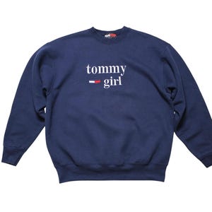 May include: Navy blue sweatshirt with the text "tommy girl" in white script. The design includes a red and white stripe below the word "tommy". The long-sleeved top is made of a soft material.