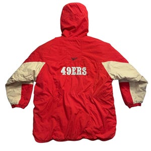 Pode incluir: Casaco com capuz vermelho e bege com o texto "49ERS" em branco. O casaco tem um corpo vermelho com mangas bege e um capuz. O logotipo da Reebok é visível acima do texto.