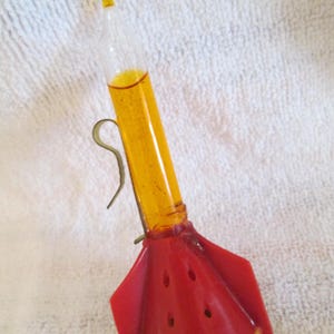May include: A rocket-shaped ornament featuring a clear glass tube filled with amber-colored liquid. The base is a bright red plastic cone with three fins and small yellow feet. A thin metal wire loop is attached to the side of the glass tube.