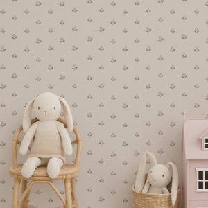 May include: A nursery scene with a beige floral wallpaper backdrop. A stuffed bunny sits on a small rattan chair. A pink dollhouse and a woven basket with another bunny are on the floor. Wooden blocks are stacked nearby.