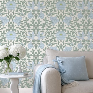 May include: A living room scene featuring a floral wallpaper in soft blue and green tones. A beige sofa with a light blue pillow and a blue throw blanket is in the foreground. A small white table holds a vase of white flowers.