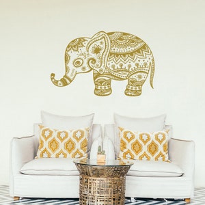 May include: A gold decorative elephant wall decal with intricate mandala patterns. The decal is shown on a white wall above a white couch with patterned throw pillows.