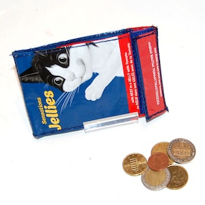 Könnte beinhalten: Eine blaue und rote Geldbörse mit einer Katze auf der Vorderseite. Die Geldbörse trägt den Text "Sensations Jellies" und "mit RUND in een SAUS mit Tomaten". Auf einer weißen Oberfläche liegen mehrere Euro-Münzen.