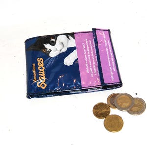 Könnte beinhalten: Eine blaue Geldbörse aus einer Katzenfuttertüte, mit einem Schwarz-Weiß-Katzenbild und dem Wort "Sensations Sauces". Mehrere Münzen sind in der Nähe verstreut. Die Geldbörse ist gefaltet und scheint handgefertigt zu sein.