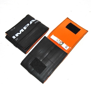 Könnte beinhalten: Schwarzes Portemonnaie aus recyceltem Reifenmaterial. Das Portemonnaie ist geöffnet und zeigt ein orangefarbenes Innenfutter und eine kleine Karte. Auf der Außenseite steht das Wort "IMPA" in weißen Buchstaben. Das andere Bild zeigt das geschlossene Portemonnaie.